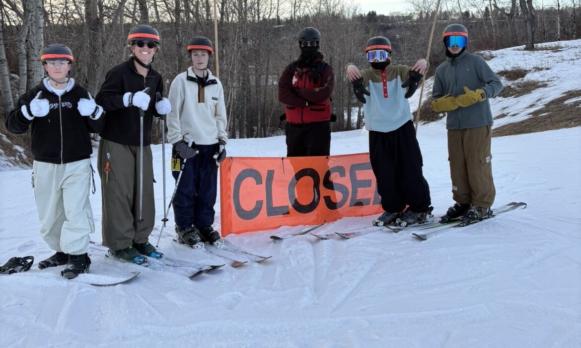 The 2-1 crew in full gear posing behind an orange CLOSED course banner on a snowy hillside